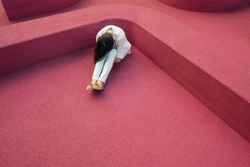 Woman sitting on vibrant pink geometric surface, head down, wearing casual attire.