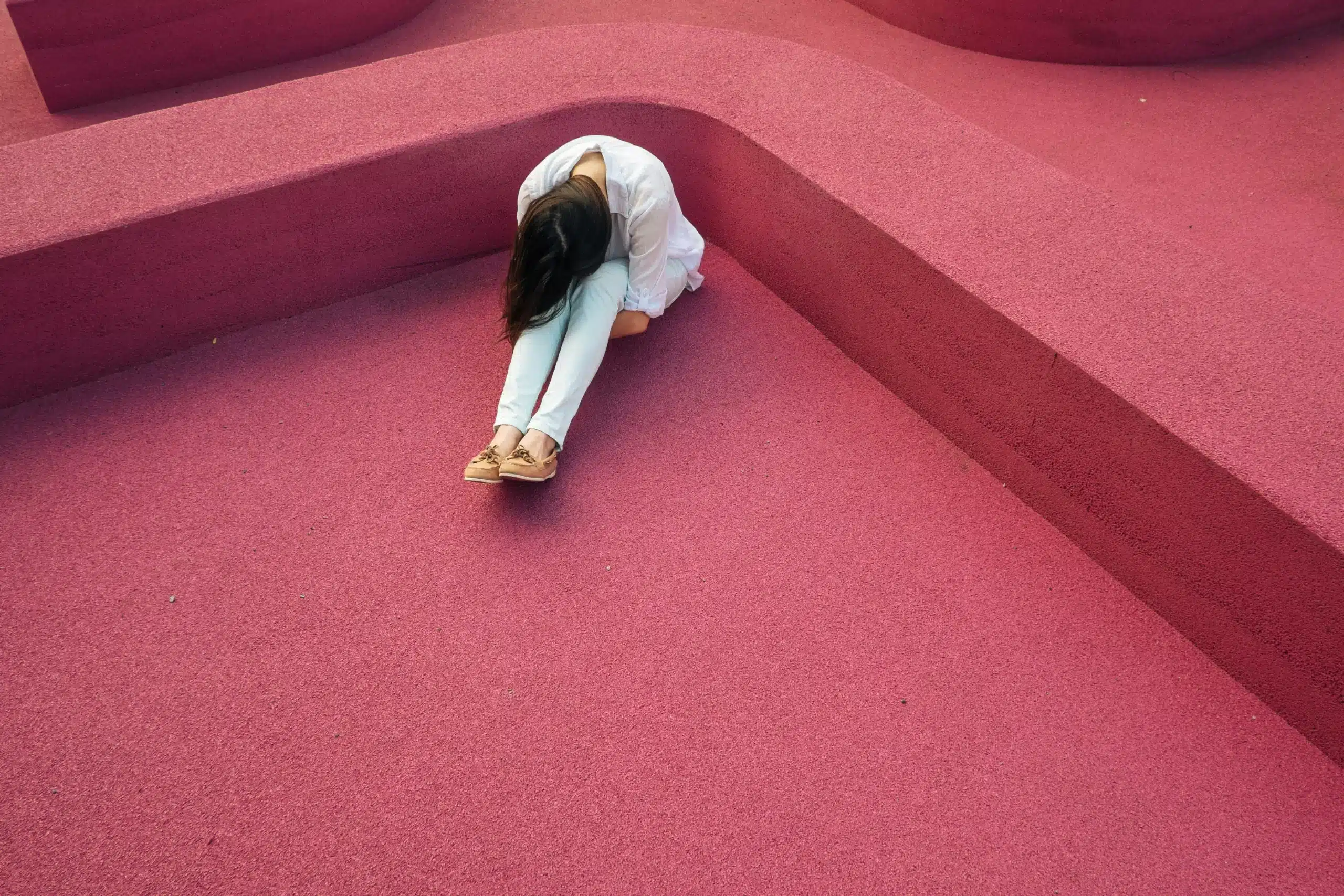 Woman sitting on vibrant pink geometric surface, head down, wearing casual attire.