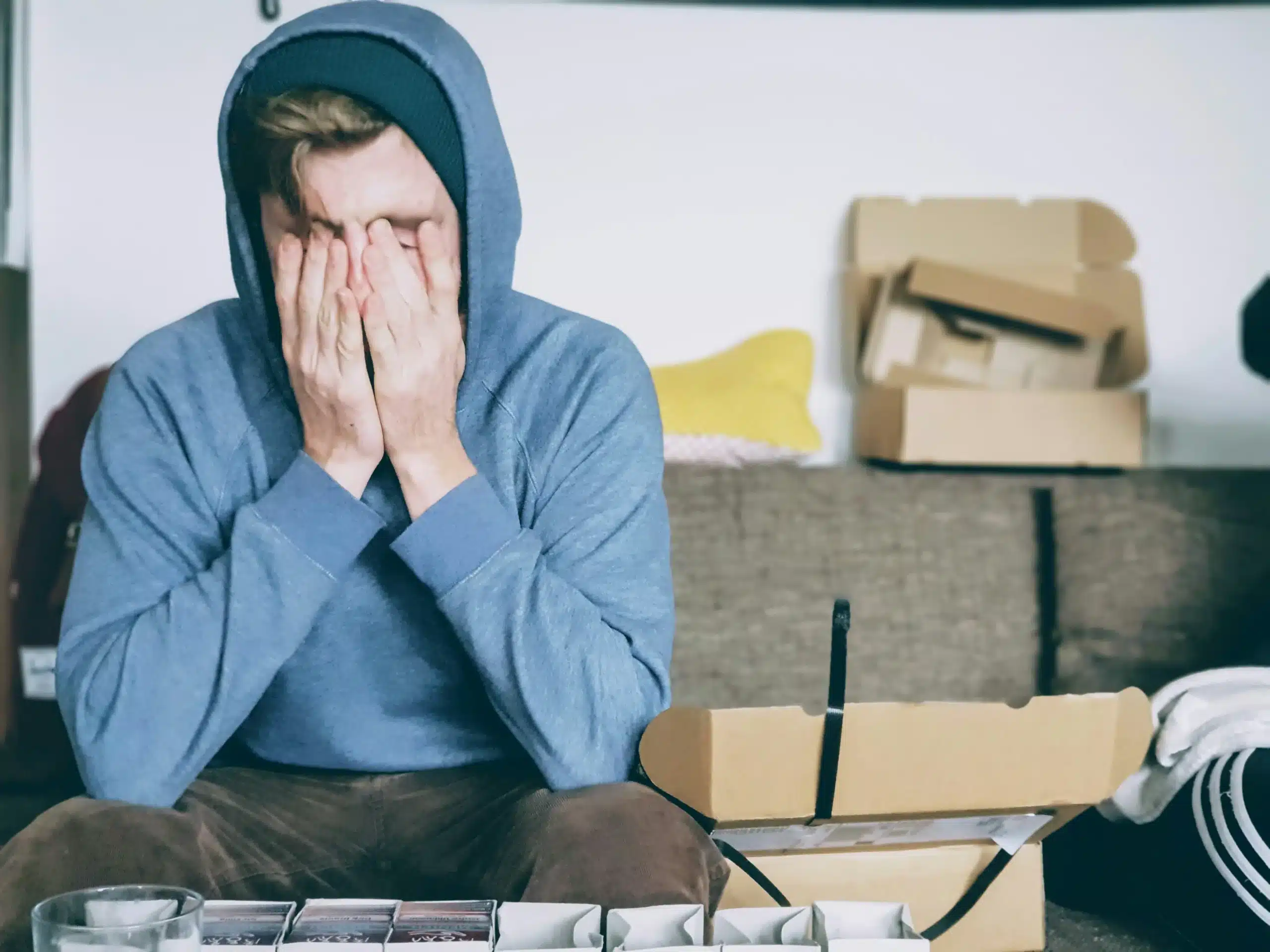 Stressed person in blue hoodie sits on a couch with face in hands, surrounded by opened cardboard boxes.